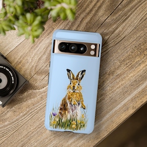 May include: A light blue phone case with a brown rabbit illustration. The rabbit is standing in a field of green grass and yellow flowers. The phone case has a black camera bump with three gold lenses.