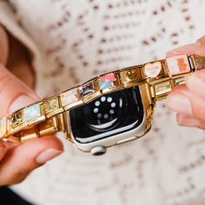 Puede incluir: Un Apple Watch dorado con una pulsera de charms colorida. La pulsera presenta pequeños charms con varios diseños, incluyendo corazones, caritas sonrientes y otros símbolos.