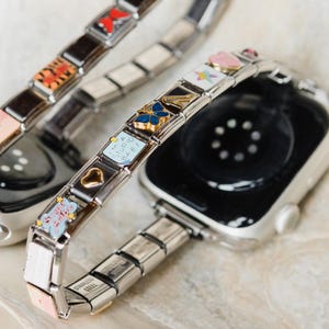 May include: Two silver watch bands with charms. The charms are colorful and include a butterfly, a heart, a star, and a small bear. The bands are made of metal links.