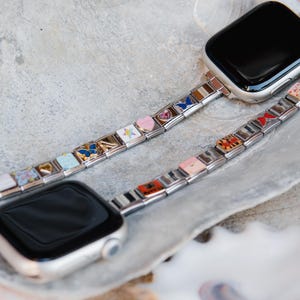 Op de afbeelding: Twee zilveren Apple Watch-bandjes met kleurrijke bedels. De bandjes zijn gemaakt van metaal en hebben een gepolijste afwerking. De bedels zijn klein en hebben verschillende ontwerpen, waaronder vlinders, harten en sterren.