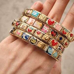 Op de afbeelding: Een gouden armband met een verscheidenheid aan kleurrijke bedels, waaronder een kat, een hart, een aardbei, een ster, een maan, een vlinder, een kers, een zon, een telefoon, een kruis en een bloem. De bedels zijn allemaal van emaille en goudkleurig.