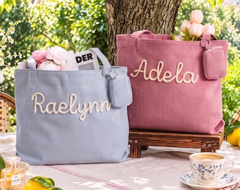 May include: Two corduroy tote bags in light blue and dusty rose, each featuring a small matching pouch attached to the strap. The blue bag displays the name Raelynn in white script, while the rose bag features the name Adela in white script.