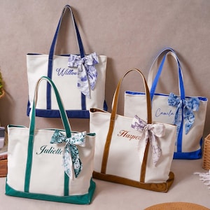 May include: Four canvas tote bags in cream with colored bases and handles in navy, green, brown, and blue. Each bag features a personalized name embroidered in cursive script and a decorative patterned ribbon tied to one of the top handles.