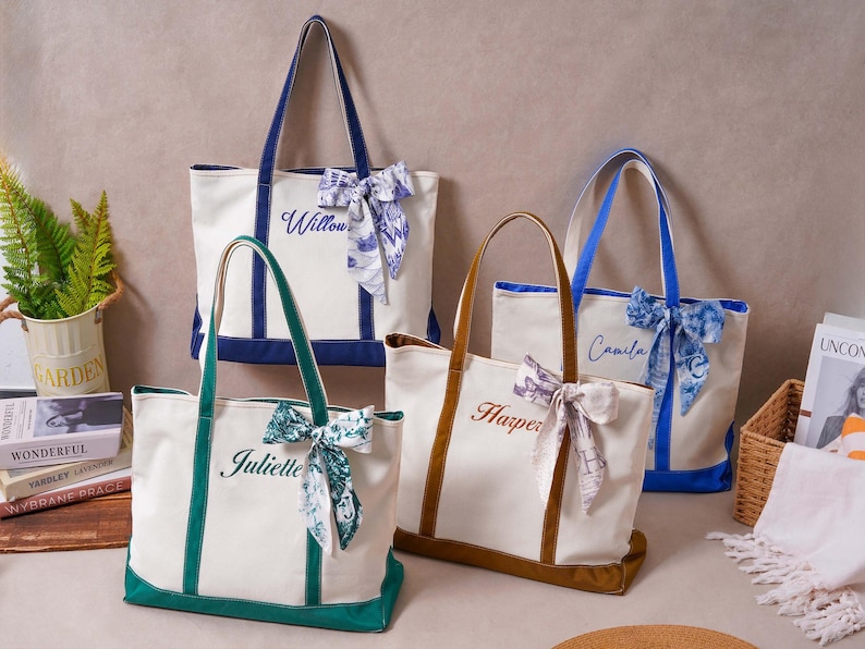 May include: Four canvas tote bags in cream with colored bases and handles in navy, green, brown, and blue. Each bag features a personalized name embroidered in cursive script and a decorative patterned ribbon tied to one of the top handles.