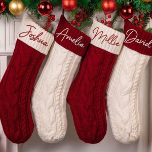 Op de afbeelding: Vier kerstsokken, twee rode en twee witte, hangen aan een schoorsteenmantel. Elke sok heeft een kabelgebreid patroon en een witte manchet met een in sierlijke letters geborduurde naam: Joshua, Amelia, Millie en David. Feestelijke decoratie.