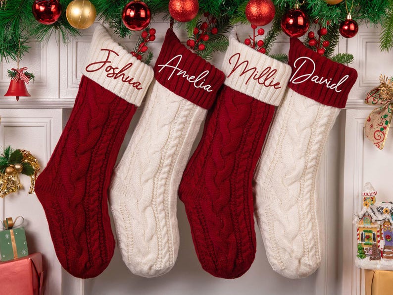 Op de afbeelding: Vier kerstsokken, twee rode en twee witte, hangen aan een schoorsteenmantel. Elke sok heeft een kabelgebreid patroon en een witte manchet met een in sierlijke letters geborduurde naam: Joshua, Amelia, Millie en David. Feestelijke decoratie.
