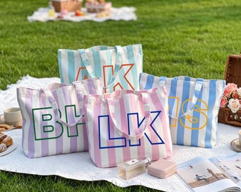 May include: Four rectangular canvas tote bags with vertical stripes in light blue, lavender, and pink. Each bag features large, bold, outlined capital letters on the front, including AK, BL, LK, and IS, with matching fabric shoulder straps.