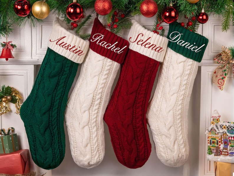 Op de afbeelding: Vier kabelgebreide kerstsokken hangen aan een schoorsteenmantel, elk met een naam op de manchet: Austin, Rachel, Selena en Daniel. De sokken zijn groen, rood en wit, met feestelijke decoraties erboven.