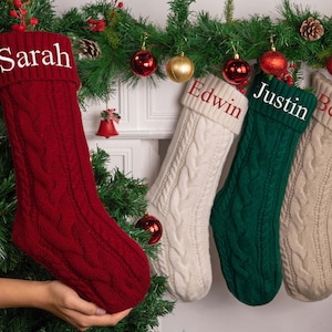 Op de afbeelding: Vier kerstsokken met kabelbreisel in rood, wit, groen en beige. Elke sok heeft een naam op de manchet: Sarah, Edwin, Justin en Bella. De sokken worden getoond met feestelijke vakantiedecoraties.
