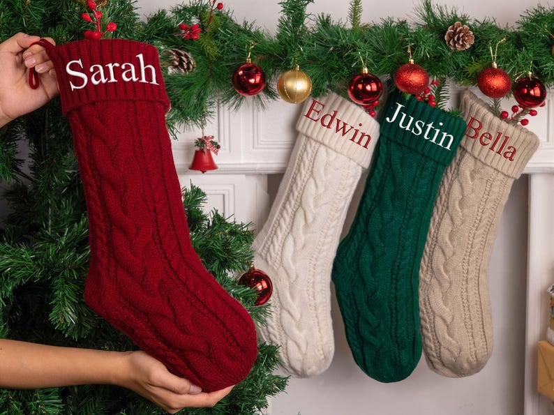 Op de afbeelding: Vier kerstsokken met kabelbreisel in rood, wit, groen en beige. Elke sok heeft een naam op de manchet: Sarah, Edwin, Justin en Bella. De sokken worden getoond met feestelijke vakantiedecoraties.