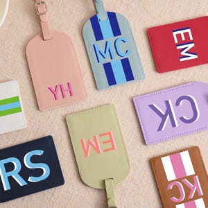 May include: A collection of rectangular luggage tags in various colors including pink, light blue, red, white, lavender, navy, olive green, and brown. Each tag features personalized two-letter monograms and striped patterns with buckle straps.