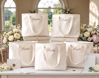 May include: Five off-white canvas tote bags with two handles each, featuring embroidered names in a script font. The names Regina, Evelyn, Jackie, Bailey, and Layla are stitched in a light brown thread on the front of each individual bag.