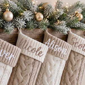 May include: A row of four knit Christmas stockings in cream and beige tones, each featuring a ribbed cuff embroidered with names including Nicole, Alison, and Scott, hanging against a rustic wooden surface with pine greenery and gold ornaments.