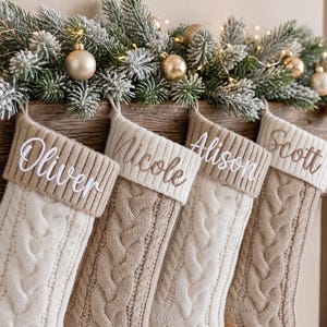 May include: Four knit Christmas stockings in cream and beige tones, each featuring a ribbed cuff embroidered with the names Oliver, Nicole, Alison, and Scott. The stockings hang from a wooden mantel decorated with frosted pine branches and gold ornaments.