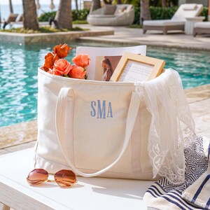 May include: A cream-colored canvas tote bag featuring the embroidered blue monogram SMA on the front. The bag contains orange flowers, a framed photograph, and a white lace fabric. A pair of brown-tinted sunglasses rests in front of the bag.