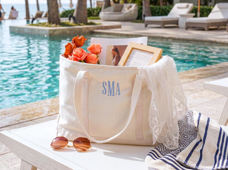 May include: A cream-colored canvas tote bag featuring the embroidered blue monogram SMA on the front. The bag contains orange flowers, a framed photograph, and a white lace fabric. A pair of brown-tinted sunglasses rests in front of the bag.