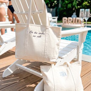 May include: Two off-white canvas tote bags. The larger bag features the black embroidered text "On the rocks, no salt" on the front. The smaller bag features the blue embroidered monogram "SMA" on the front. Both bags have two shoulder straps.