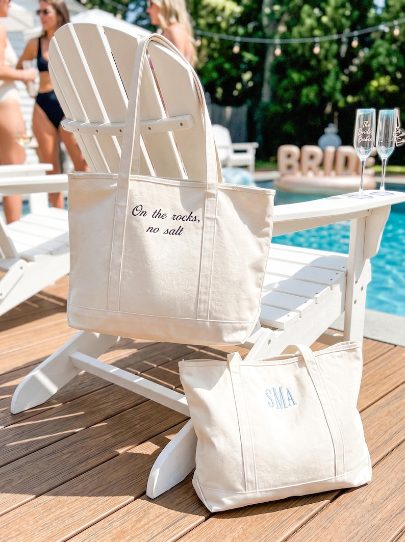 May include: Two off-white canvas tote bags. The larger bag features the black embroidered text "On the rocks, no salt" on the front. The smaller bag features the blue embroidered monogram "SMA" on the front. Both bags have two shoulder straps.