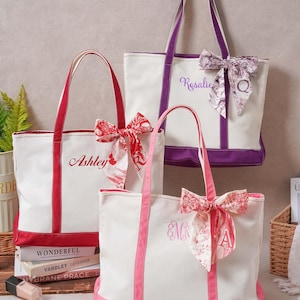 May include: Three canvas tote bags in white with red, purple, and pink trim. Each bag features a matching fabric bow tied to the handle, personalized embroidery, and a letter. The bags are labeled with the names Ashley, Rosalie, and EMR.