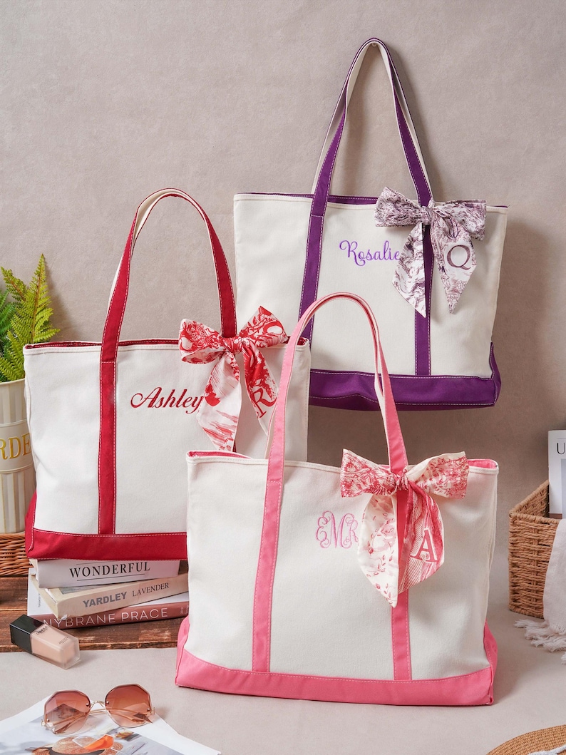 May include: Three canvas tote bags in white with red, purple, and pink trim. Each bag features a matching fabric bow tied to the handle, personalized embroidery, and a letter. The bags are labeled with the names Ashley, Rosalie, and EMR.