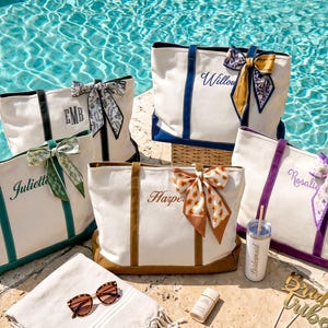 May include: Five off-white canvas tote bags with colored bases and handles, each featuring a personalized name or monogram and a decorative patterned ribbon tied to one handle. A tumbler, sunglasses, and a sign are placed in the foreground.