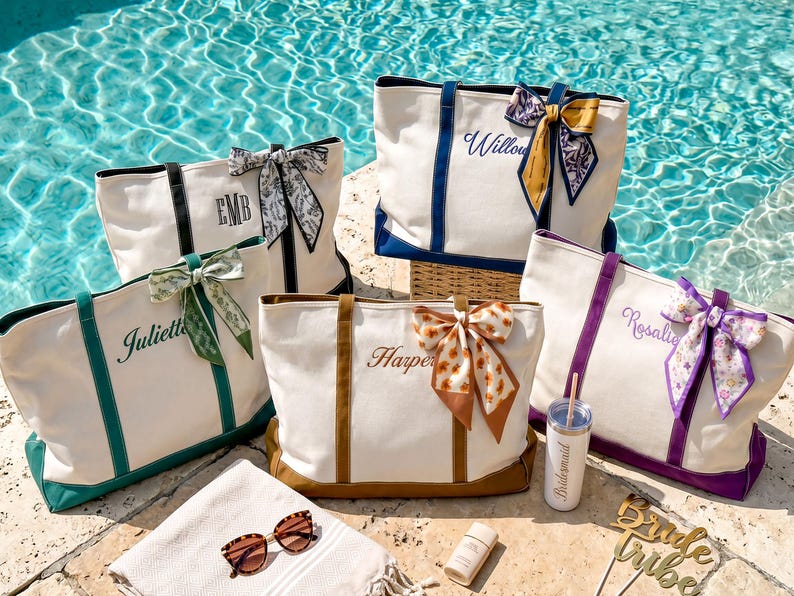 May include: Five off-white canvas tote bags with colored bases and handles, each featuring a personalized name or monogram and a decorative patterned ribbon tied to one handle. A tumbler, sunglasses, and a sign are placed in the foreground.