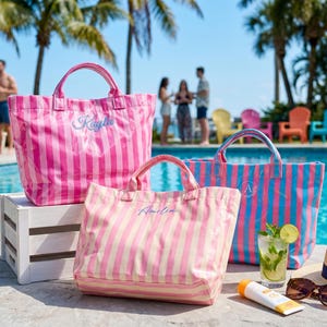 May include: Three striped tote bags in pink, light pink, and blue, each with a name embroidered on the front. The bags are near a pool with a drink, sunglasses, and a sun hat. Palm trees and people are in the background.