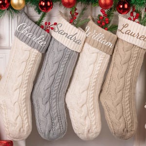 Op de afbeelding: Vier kabelgebreide kerstsokken in cr&egrave;me, grijs en beige hangen aan een slinger. Elke sok heeft een contrasterende manchet met een witte naam: Charlie, Sandra, William en Laurel. Feestelijke vakantiedecoratie omringt de sokken.