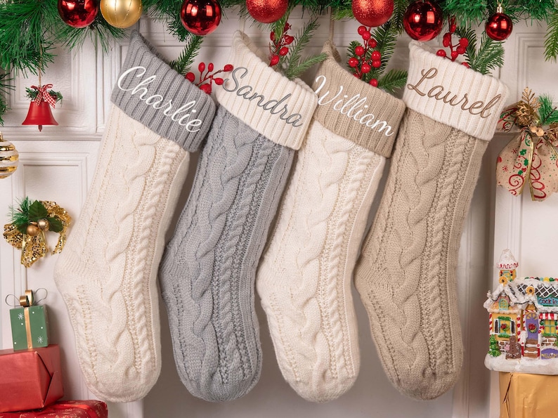 Op de afbeelding: Vier kabelgebreide kerstsokken in cr&egrave;me, grijs en beige hangen aan een slinger. Elke sok heeft een contrasterende manchet met een witte naam: Charlie, Sandra, William en Laurel. Feestelijke vakantiedecoratie omringt de sokken.