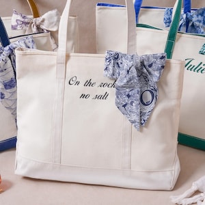May include: A cream-colored canvas tote bag featuring the black embroidered text On the rocks no salt and a blue and white patterned fabric bow tied to the handle. Other similar tote bags with different colored accents are visible nearby.