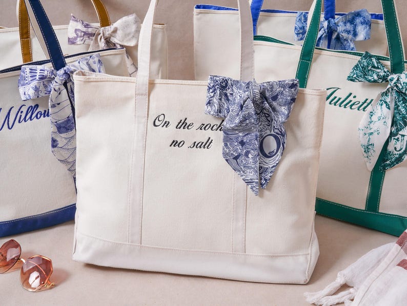 May include: A cream-colored canvas tote bag featuring the black embroidered text On the rocks no salt and a blue and white patterned fabric bow tied to the handle. Other similar tote bags with different colored accents are visible nearby.