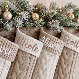 May include: A row of four knitted Christmas stockings in cream and beige tones, each featuring a ribbed cuff embroidered with the names Nicole, Alison, and Scott. The stockings have a cable knit pattern and hang from a wooden mantelpiece.
