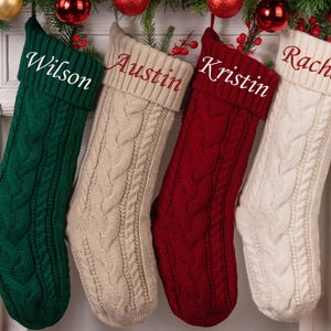 Op de afbeelding: Vier kabelgebreide kerstsokken in groen, beige, rood en wit, elk met een naam geborduurd op de manchet: Wilson, Austin, Kristin en Rachel. De sokken hangen aan een slinger met rode ornamenten.