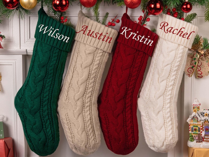Op de afbeelding: Vier kabelgebreide kerstsokken in groen, beige, rood en wit, elk met een naam geborduurd op de manchet: Wilson, Austin, Kristin en Rachel. De sokken hangen aan een slinger met rode ornamenten.