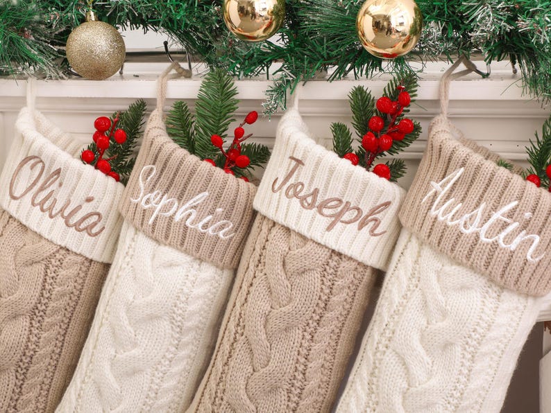 Op de afbeelding: Vier gepersonaliseerde kerstsokken in cr&egrave;me en tan kabelbreisel met geborduurde namen: Olivia, Sophia, Joseph en Austin. Elke sok is versierd met rode bessen en dennentakjes, met gouden ornamenten en groen in de achtergrond.