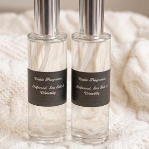 Può includere: Due flaconi spray in vetro trasparente con tappi argentati. Ogni flacone ha un'etichetta nera con il testo "Battle Fragrance Driftwood, Sea Salt & Waterlily". I flaconi sono su una superficie bianca strutturata.