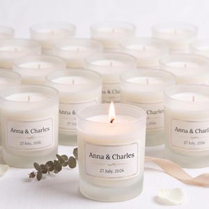 May include: A display of white candles in frosted glass holders, some lit, with labels reading "Anna & Charles" and "27 July, 2026". The candles are arranged with floral and ribbon accents, creating a soft, romantic ambiance.