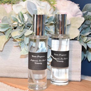 Könnte beinhalten: Zwei transparente Glassprühflaschen mit silbernen Kappen und schwarzen Etiketten. Auf den Etiketten steht "Battle Fragrance Driftwood, Sea Salt & Waterlily". Sie stehen vor einem Blumengesteck mit weißen und grünen Blättern.