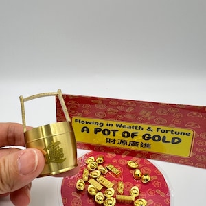 Könnte beinhalten: Ein goldfarbener Topf mit Henkel, mit chinesischen Schriftzeichen, wird vor einem rot-goldenen Hintergrund gehalten. Der Text "Flowing in Wealth & Fortune A POT OF GOLD" ist sichtbar. Kleine Goldbarren und Münzen sind auf einer roten Oberfläche verstreut.