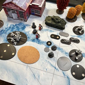 Pode incluir: Uma cena de jogo de guerra em miniatura com um tanque verde, várias figuras e um edifício com a etiqueta "Garage Automobile". A cena está ambientada em uma superfície com um padrão azul e branco com várias bases redondas e ovaladas.