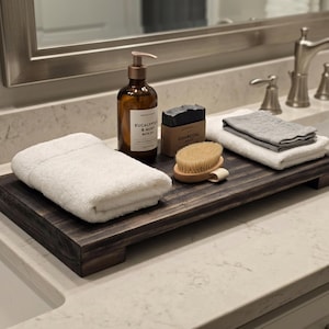 May include: A rectangular dark brown wooden tray holding a white towel, a brown glass pump bottle labeled Eucalyptus & Mint, a black charcoal soap bar, a round wooden scrub brush, and a folded grey cloth towel arranged on a white countertop.