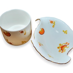 May include: A white ceramic teacup and leaf-shaped dish set. The cup and dish are decorated with orange and yellow floral patterns and gold trim. The set is designed for serving tea or small snacks.