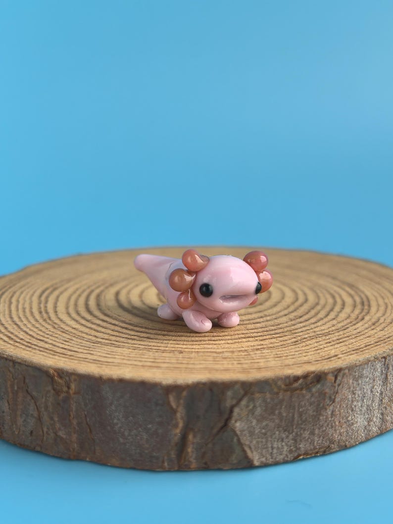 Op de afbeelding: Een kleine, roze axolotl-figuur met bruine kieuwen en zwarte ogen. Het staat op een houten schijf tegen een helderblauwe achtergrond. De axolotl is een schattig, miniatuurbeeldje.