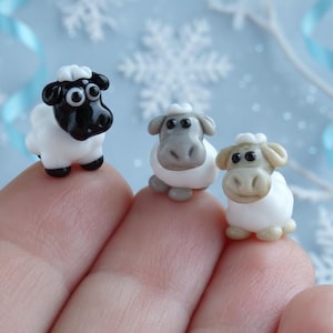 May include: Three miniature sheep figurines. One sheep has a black head and body accents, while the other two have gray and tan heads. All have white bodies and are posed on a finger. The background is a light blue with snowflake decorations.