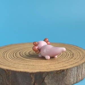 Op de afbeelding: Een kleine, roze glazen axolotl-figuur met bruine kieuwen, liggend op een houten schijf tegen een blauwe achtergrond. De axolotl heeft een gladde, glanzende afwerking en is een grillig decoratief item.