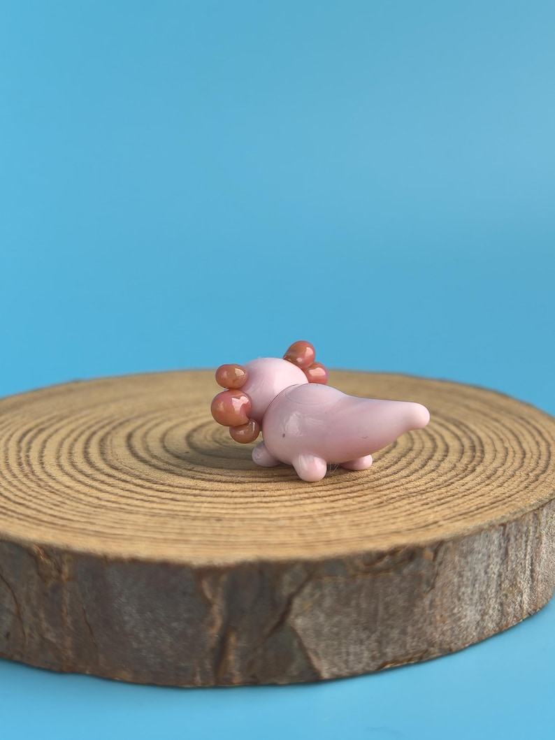 Op de afbeelding: Een kleine, roze glazen axolotl-figuur met bruine kieuwen, liggend op een houten schijf tegen een blauwe achtergrond. De axolotl heeft een gladde, glanzende afwerking en is een grillig decoratief item.