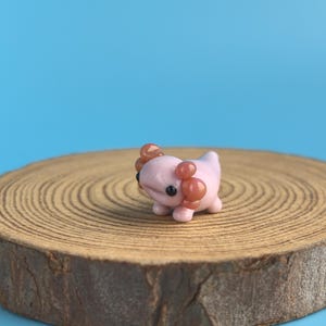 Op de afbeelding: Een kleine, roze axolotl-figuur met bruine kieuwen en zwarte ogen. De figuur staat op een ronde houten schijf met groeiringen, tegen een blauwe achtergrond. De axolotl is een handgemaakt item. De houten schijf is ongeveer 10 cm in diameter.