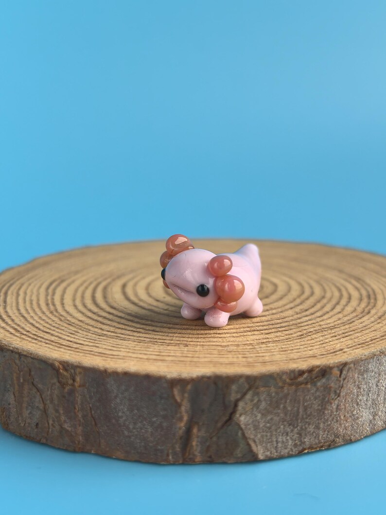 Op de afbeelding: Een kleine, roze axolotl-figuur met bruine kieuwen en zwarte ogen. De figuur staat op een ronde houten schijf met groeiringen, tegen een blauwe achtergrond. De axolotl is een handgemaakt item. De houten schijf is ongeveer 10 cm in diameter.