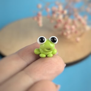 May include: A small, lime green frog figurine with large, black-pupiled eyes. The frog is perched on a finger, with a blurred blue and tan background. The frog has a smiling expression and rounded features.