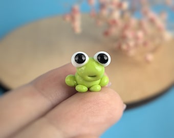 May include: A small, lime green frog figurine with large, black-pupiled eyes. The frog is perched on a finger, with a blurred blue and tan background. The frog has a smiling expression and rounded features.
